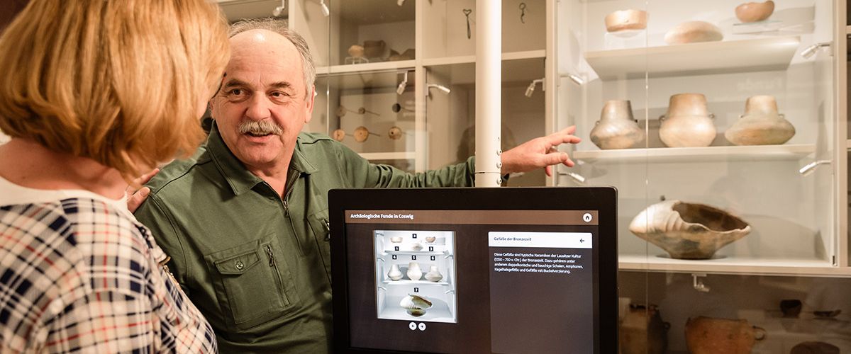 Altere Besucher stehen am Tablet und reden über Schaudepot im Raum Archäologie